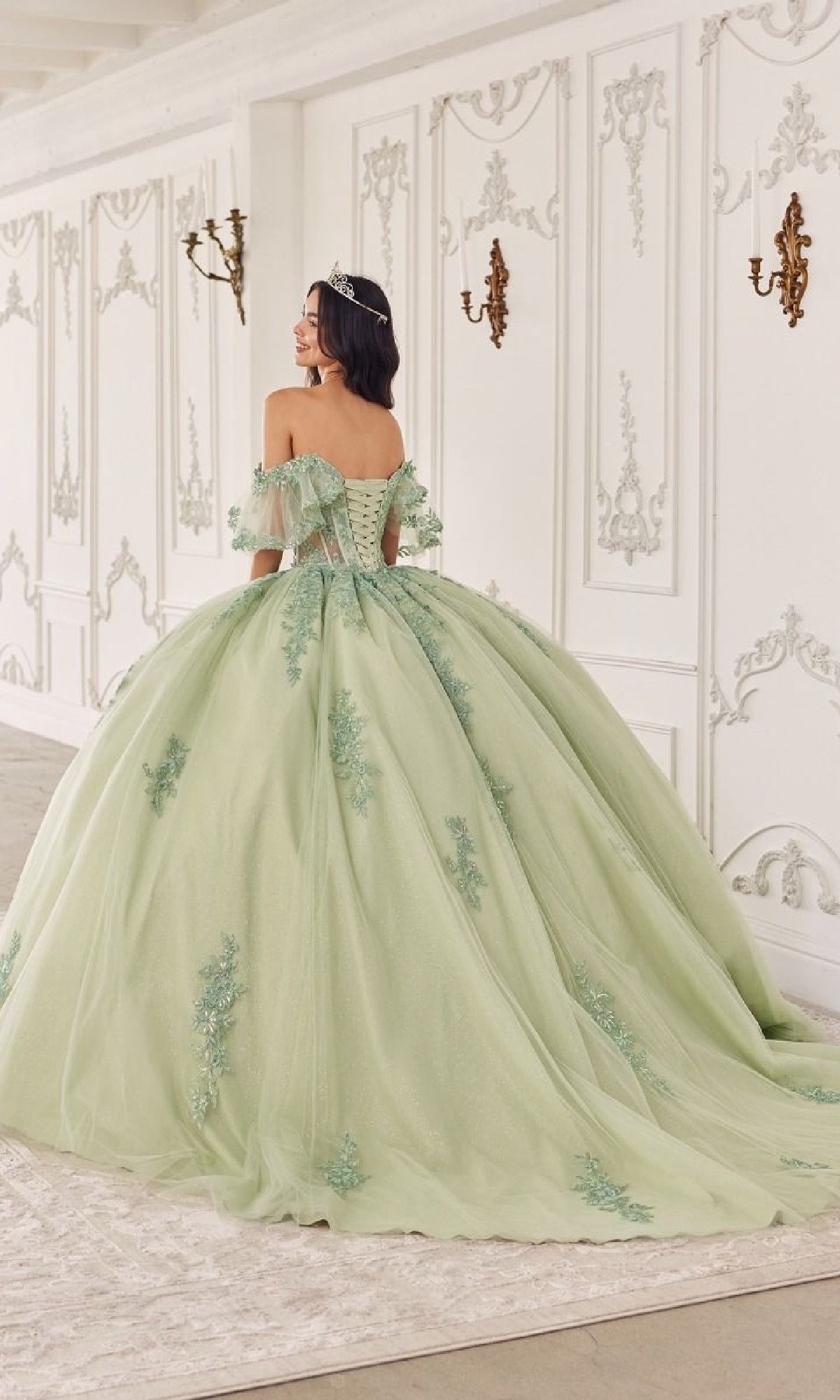 A woman in the Quinceanera Dress 15725 by Ladivine, featuring sage green floral lace appliques, stands in an ornate white room with gold wall sconces, facing away from the camera.